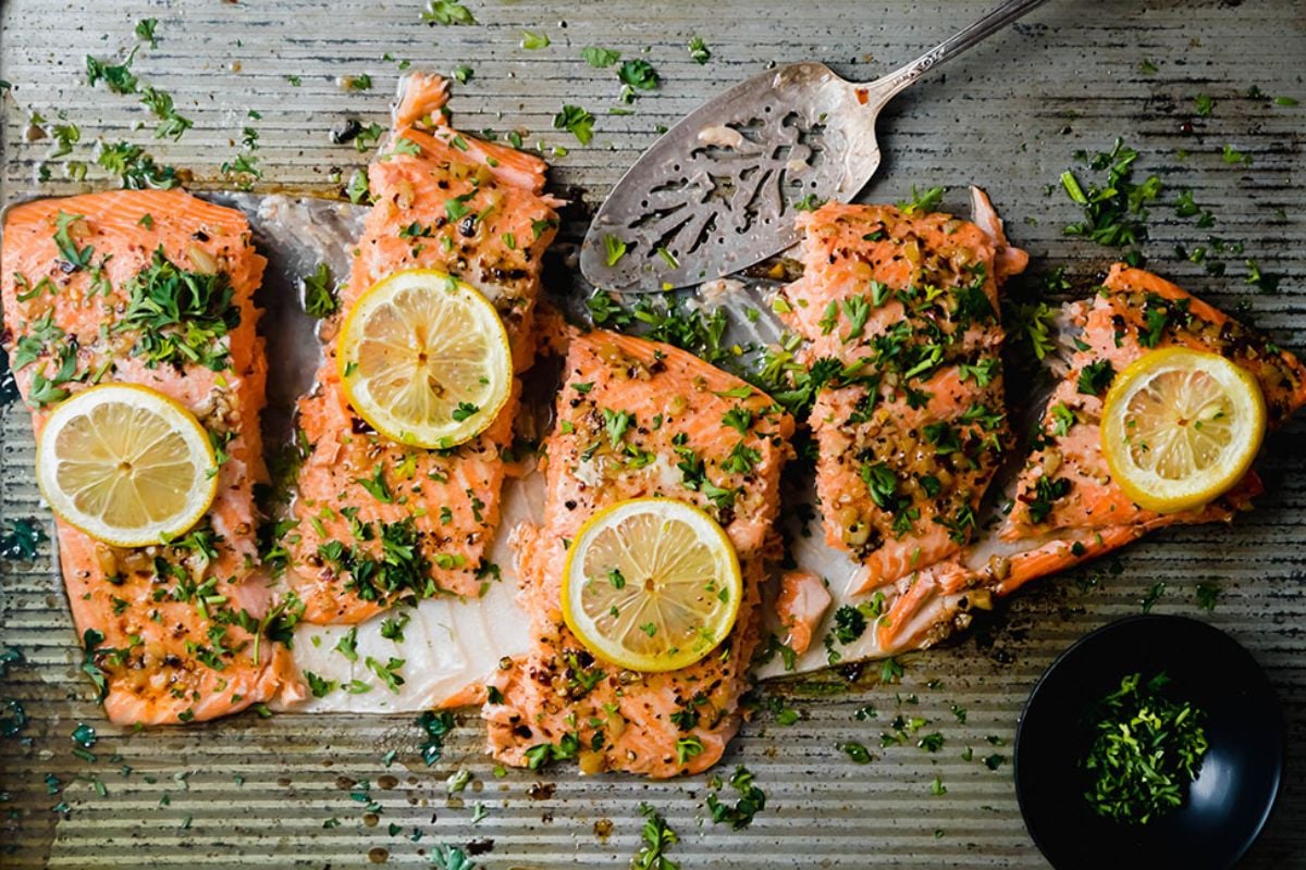 Four grilled salmon fillets garnished with lemon slices and chopped parsley on a metal baking tray, served with a small bowl of herbs. This is an example of seafood recipes.