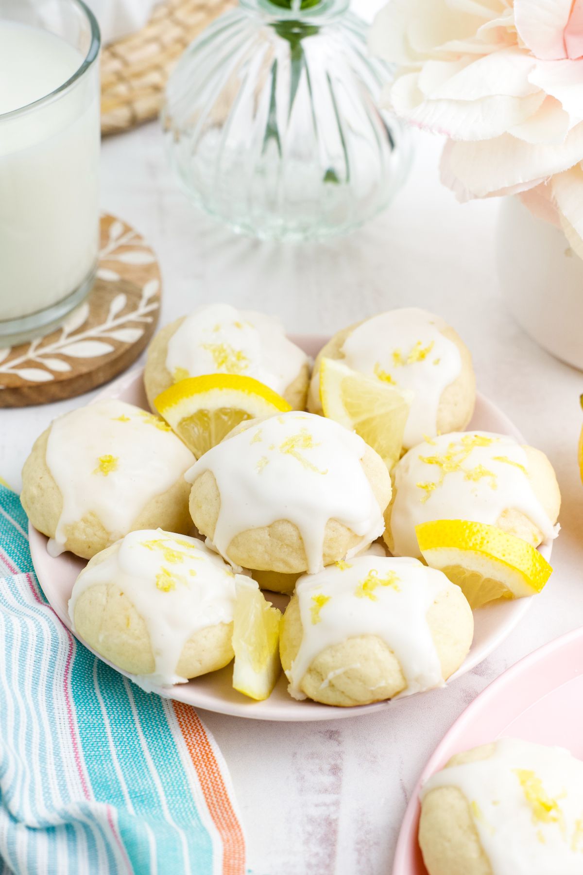 A plate of lemon ricotta cookies with white icing and lemon wedges, set beside a glass of milk and fresh flowers.