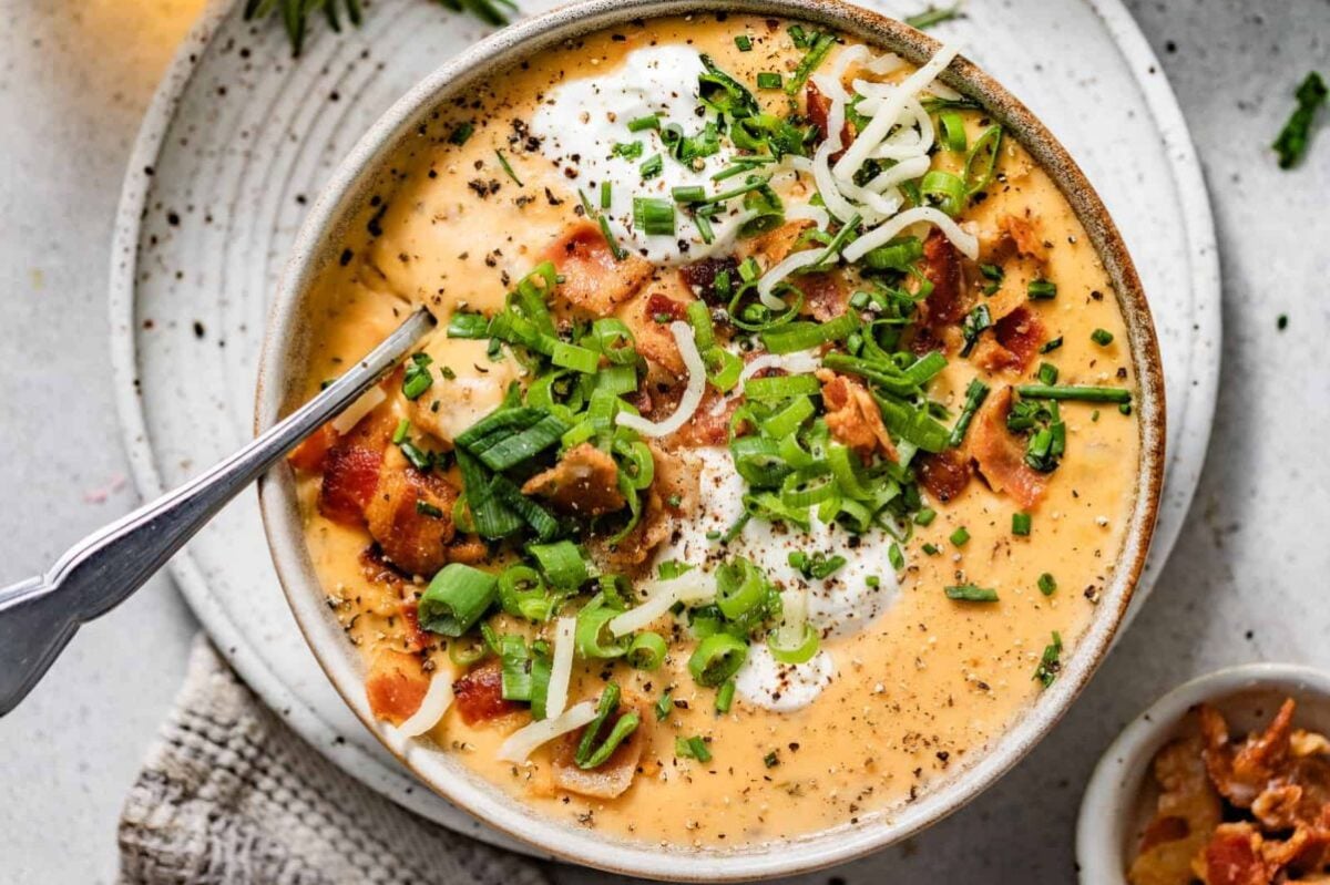Close-up of a bowl of creamy soup topped with bacon bits, shredded cheese, green onions, sour cream, and pepper. A spoon is placed in the bowl, and a small dish with toppings is visible on the side.