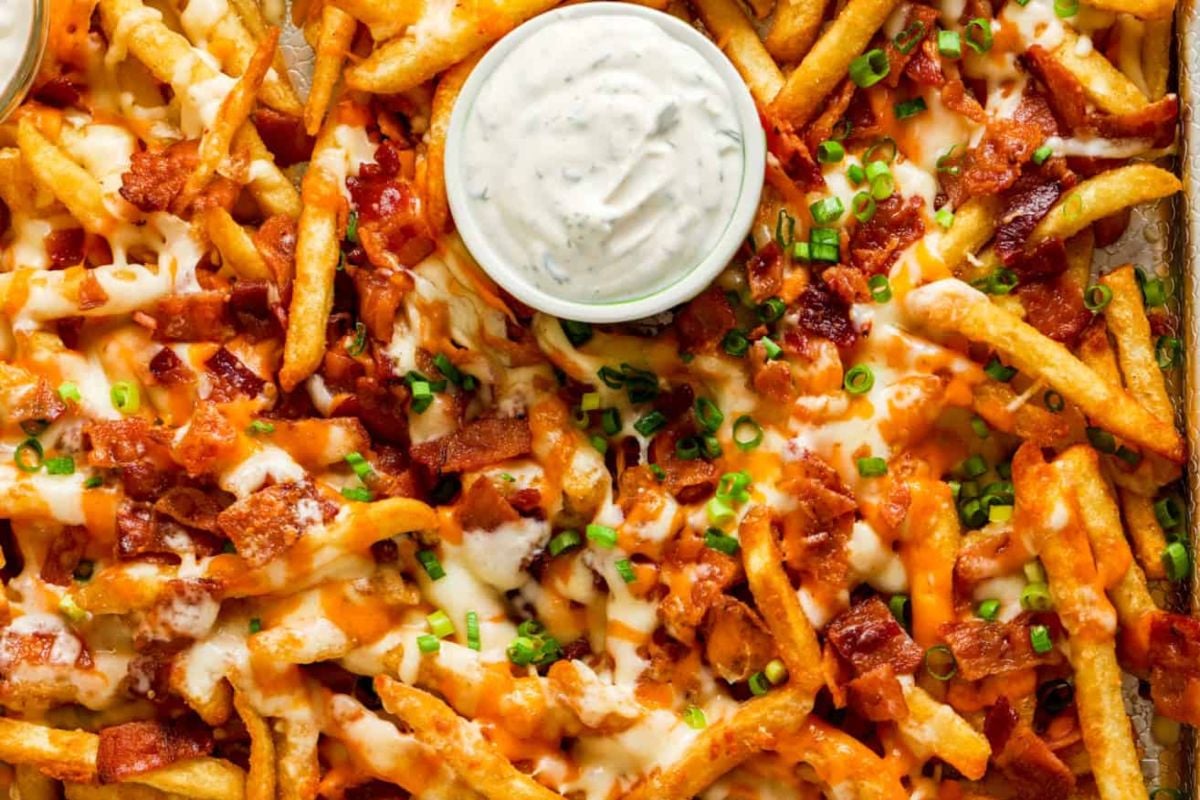A plate of cheesy loaded fries topped with melted cheese, bacon bits, and green onions, served with a side of ranch dressing.