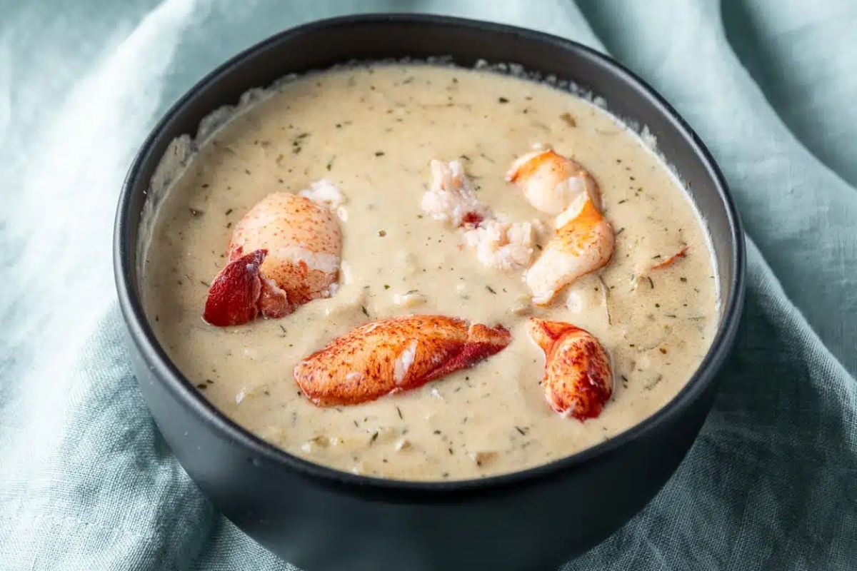 A bowl of creamy lobster bisque garnished with chunks of lobster meat, served on a textured blue cloth, perfect for seafood recipes.