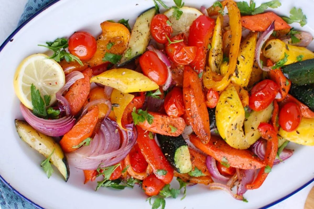 A plate of roasted Easter side vegetables garnished with fresh herbs and lemon wedges.