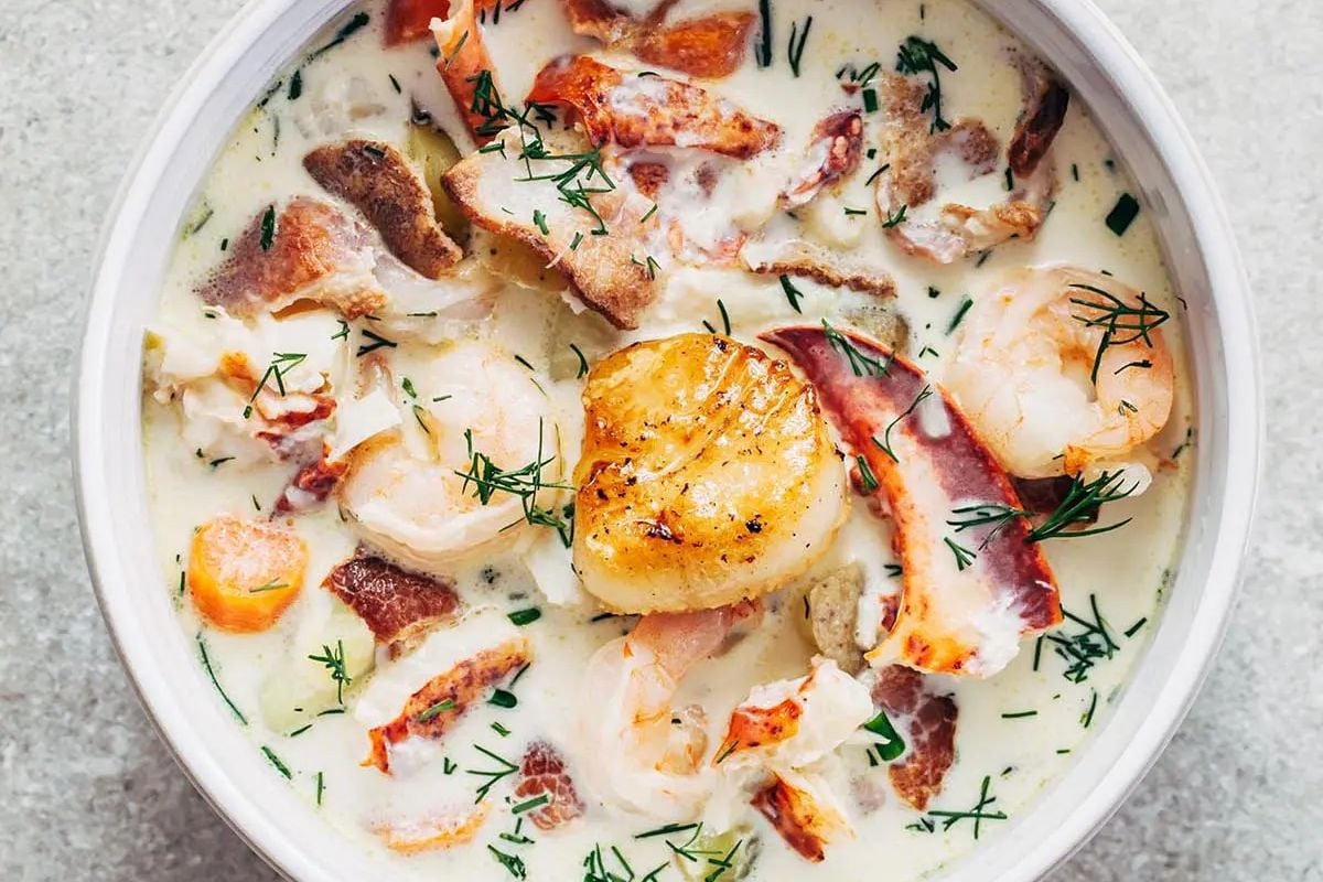 Creamy seafood chowder with shrimp, scallop, bacon, herbs, and lobster pieces in a white bowl—a comforting classic for fans of delicious seafood recipes.