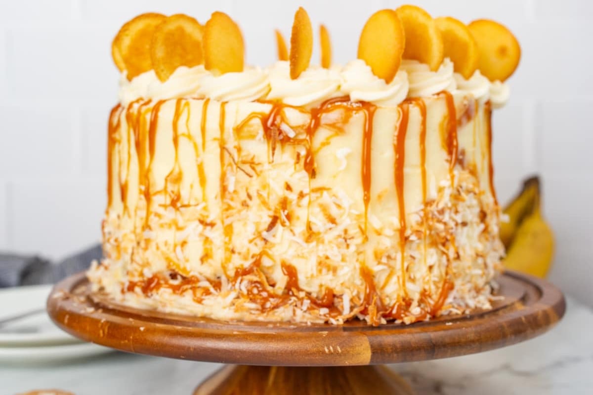 Old fashioned layered coconut cake with caramel drizzle and fruit topping on a wooden cake stand.