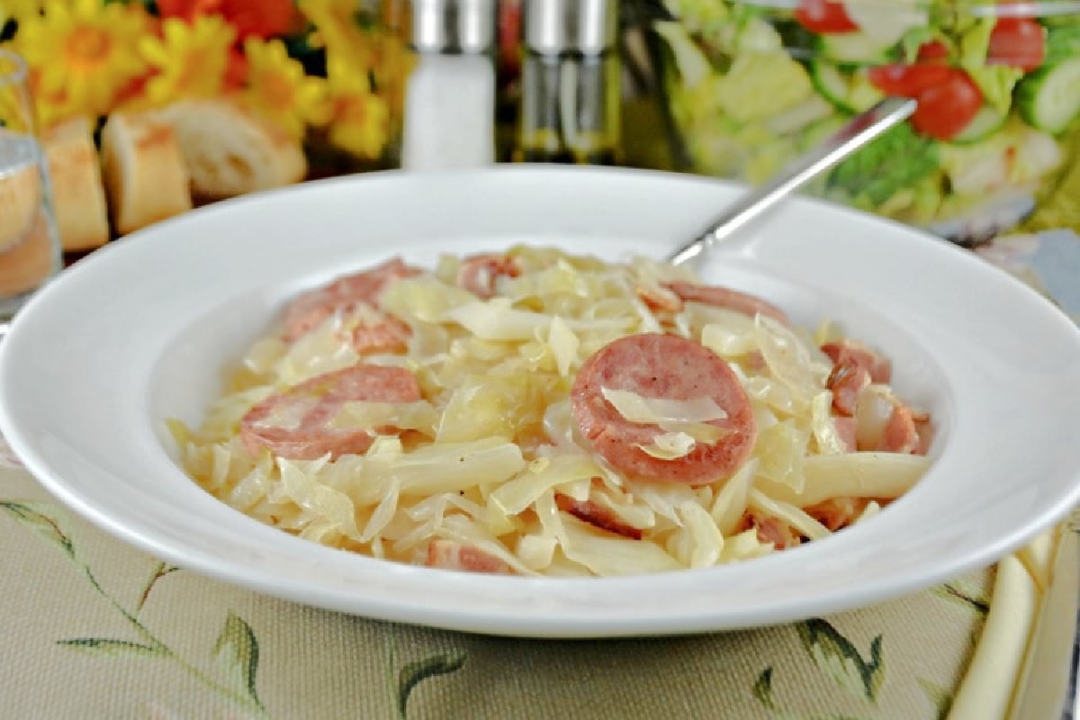 A bowl of old-fashioned pasta with sliced sausage and a creamy sauce, served on a table with condiments in the background.