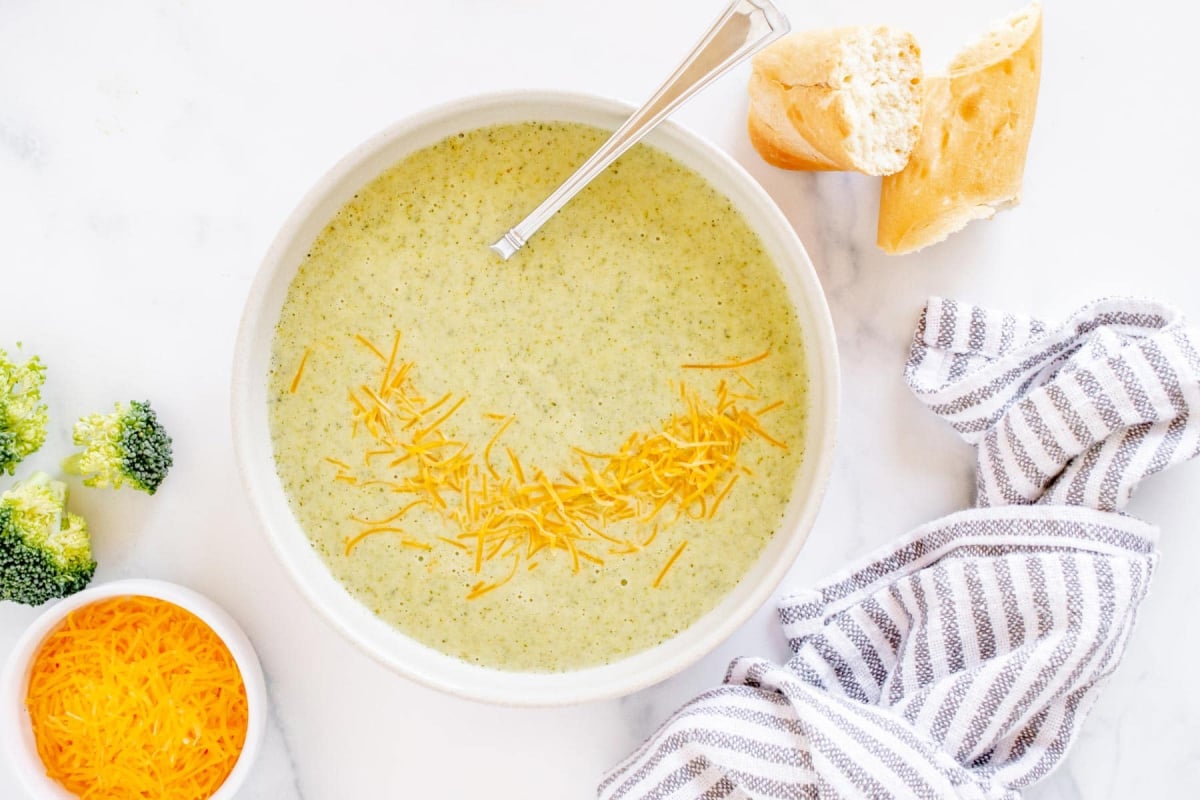 Bowl of broccoli cheddar soup topped with shredded cheese, with bread and a striped towel beside it—a cozy classic inspired by Old Fashioned Recipes.