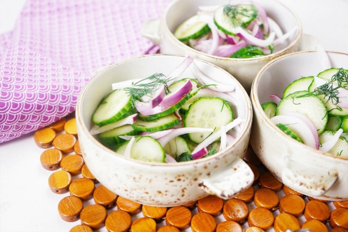 Bowls of old-fashioned cucumber salad with dill and red onion on a table with scattered wooden tokens.