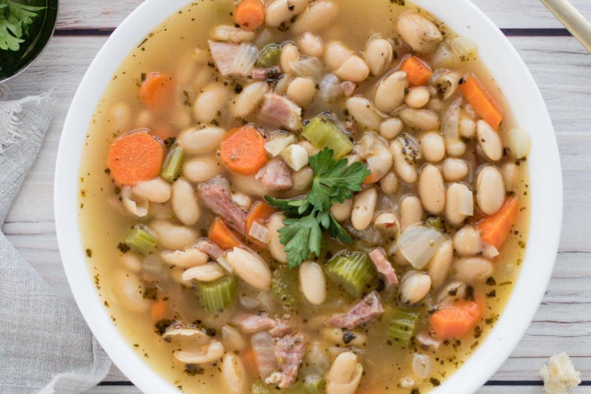 A bowl of old-fashioned bean soup with diced carrots, celery, and ham, garnished with parsley.