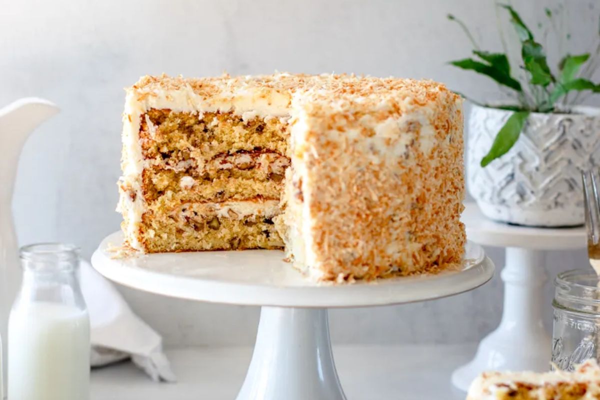 A layered carrot layer cake with cream cheese frosting and a coating of chopped nuts on a cake stand, accompanied by two bottles of milk.