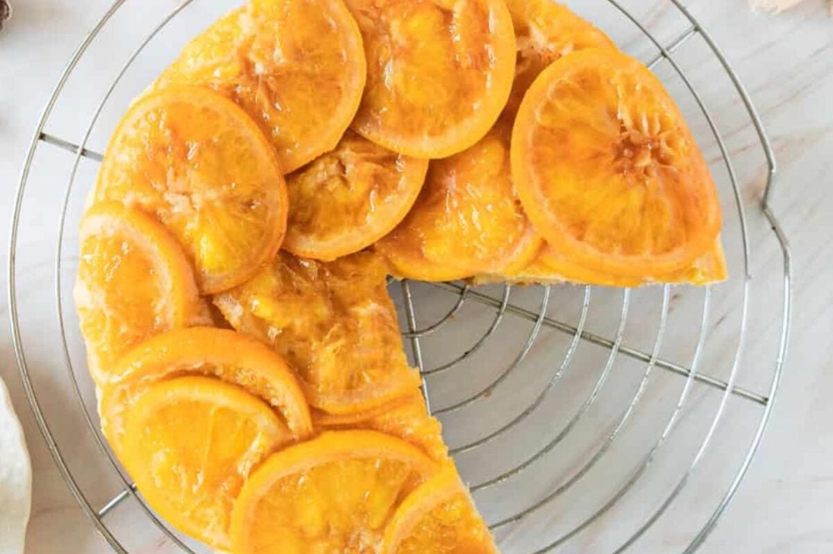 Round upside down cake topped with glazed orange slices on a cooling rack, with a large wedge missing.