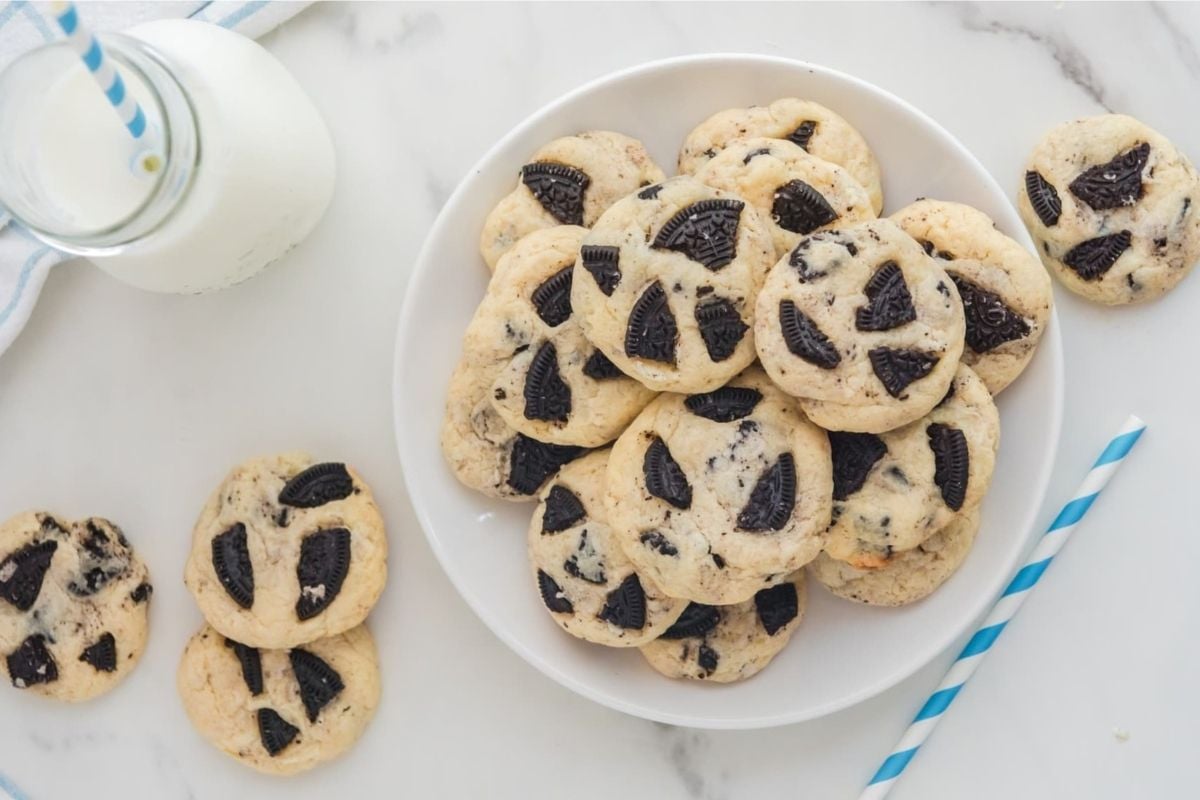 A plate of homemade cookies from our cookie recipes with chocolate sandwich cookie pieces, accompanied by a glass of milk with a striped straw.