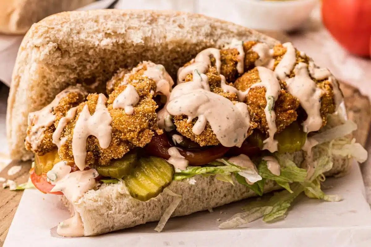 A fried seafood sandwich with lettuce, tomatoes, pickles, and drizzled with creamy sauce in a split bun.