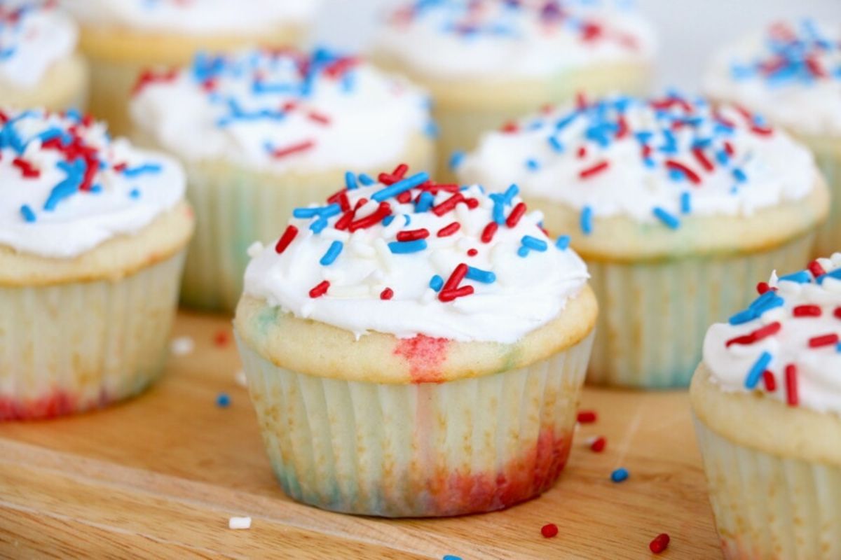 Vanilla poke cupcakes with white frosting and red and blue sprinkles on a wooden cutting board.