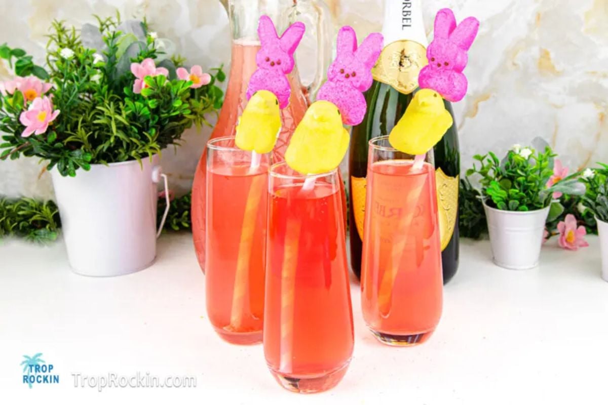 Three festive Easter glasses with a pink beverage garnished with peeps candy and a citrus slice, set against a celebratory background with a bottle of champagne.
