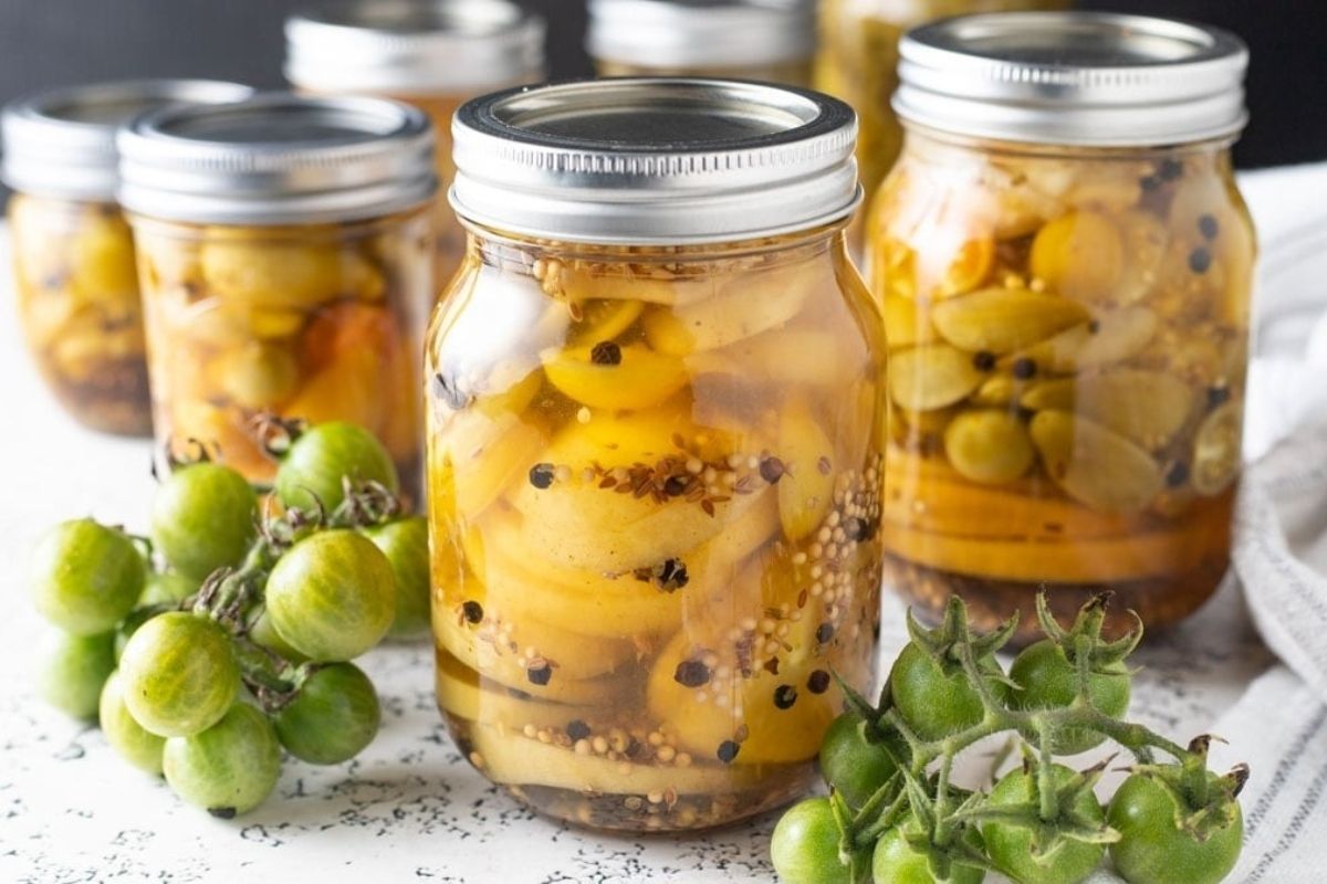 Jars of homemade pickled recipes with spices and green tomatoes nearby.