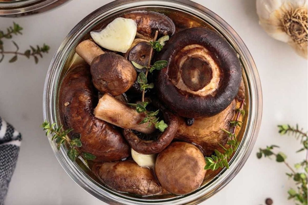 A variety of pickled mushrooms garnished with herbs in a glass jar, viewed from above.