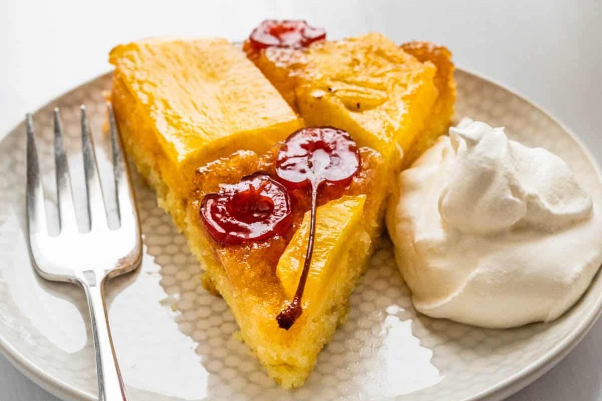 Two slices of classic upside down cakes with cherries and a dollop of whipped cream sit on a plate with a fork.