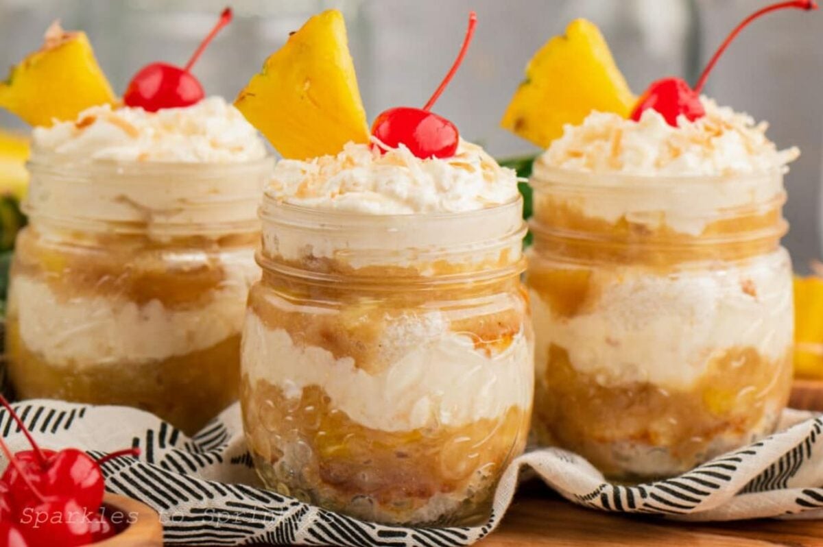 Three jars of single-serve individual desserts, each filled with pineapple, topped with whipped cream, shredded coconut, a cherry, and a pineapple wedge.
