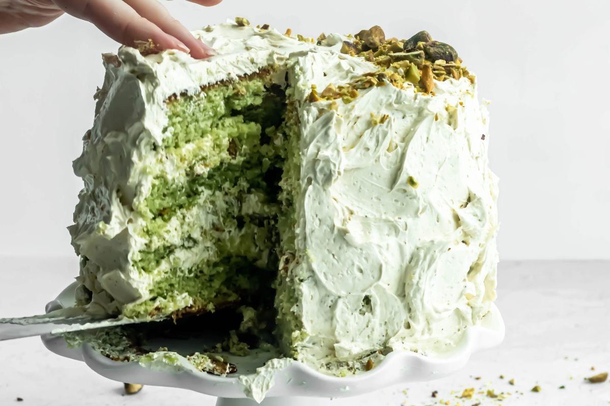 A hand serving a slice of green pistachio layer cake with white frosting on a cake stand.