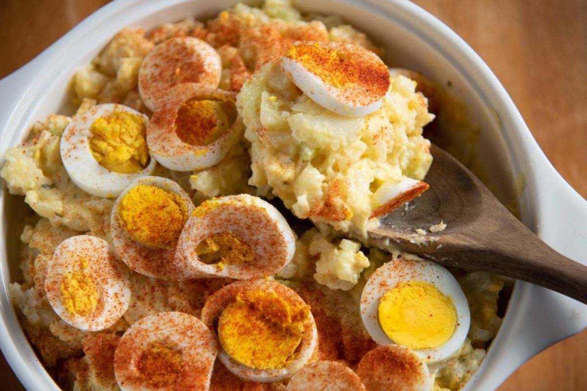 Potato salad topped with sliced hard-boiled eggs and sprinkled with paprika, served in a white dish with a wooden spoon—perfect for Easter sides.