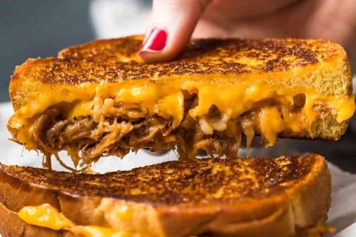 A hand holding a grilled cheese sandwich from cheesy recipes, with melted cheese and shredded meat oozing from the middle.