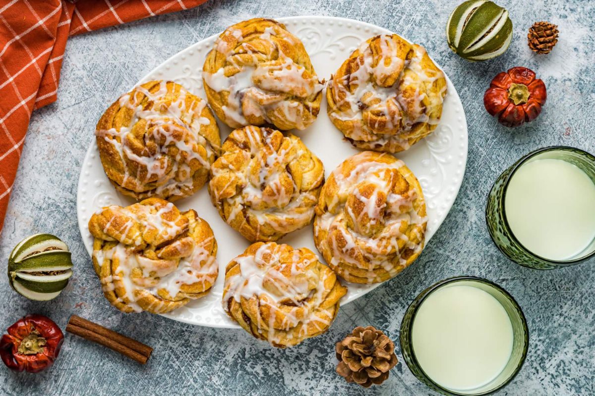 A plate of glazed cinnamon rolls with glasses of milk beside a pie recipes book and autumnal decorations on a textured surface.