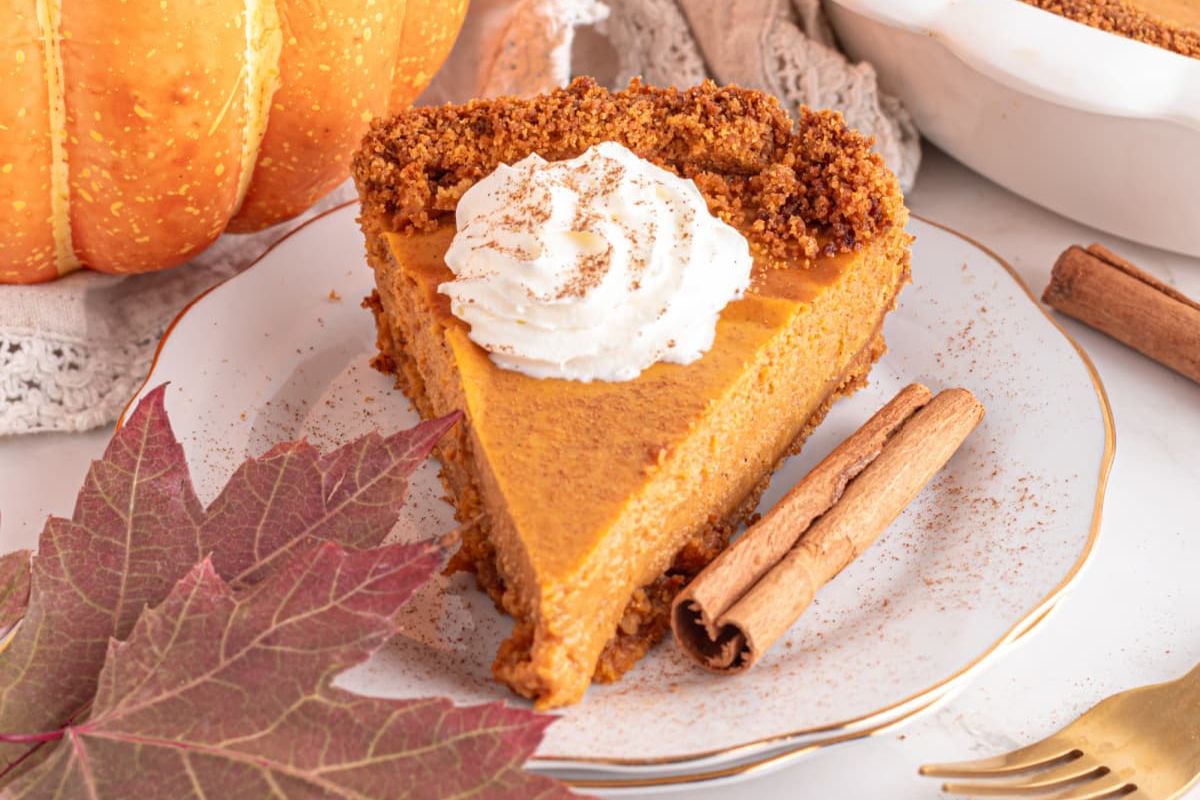 A slice of pumpkin pie from our Pie Recipes, topped with whipped cream, garnished with cinnamon sticks, adjacent to autumn leaves.
