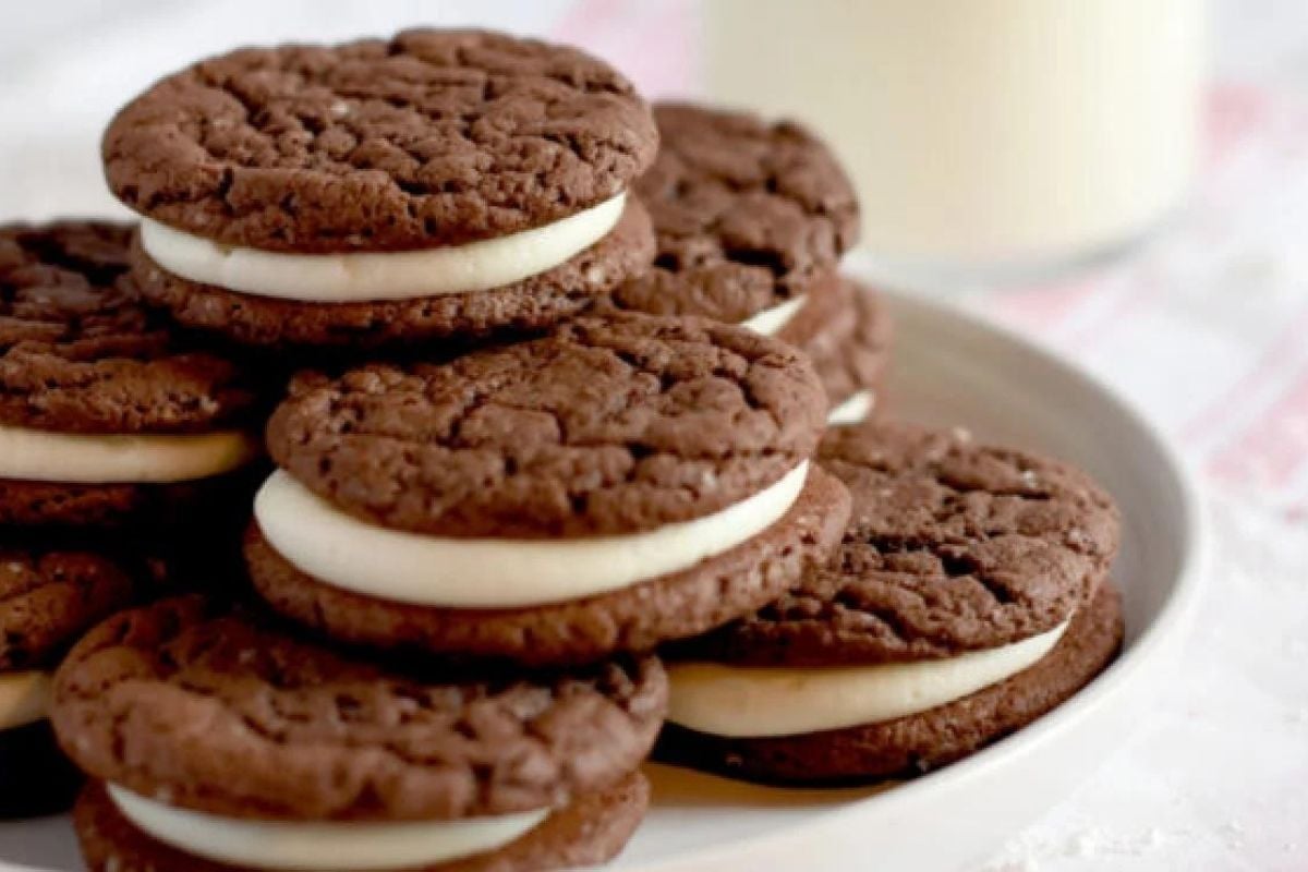 A plate of chocolate sandwich cookies with cream filling, from our top cookie recipes, is accompanied by a glass of milk in the background.