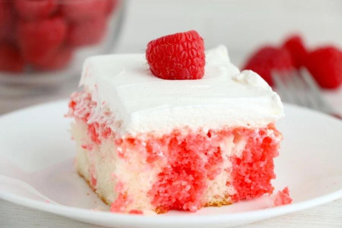 A slice of pink raspberry Poke Cake with a layer of white frosting and a single raspberry on top.