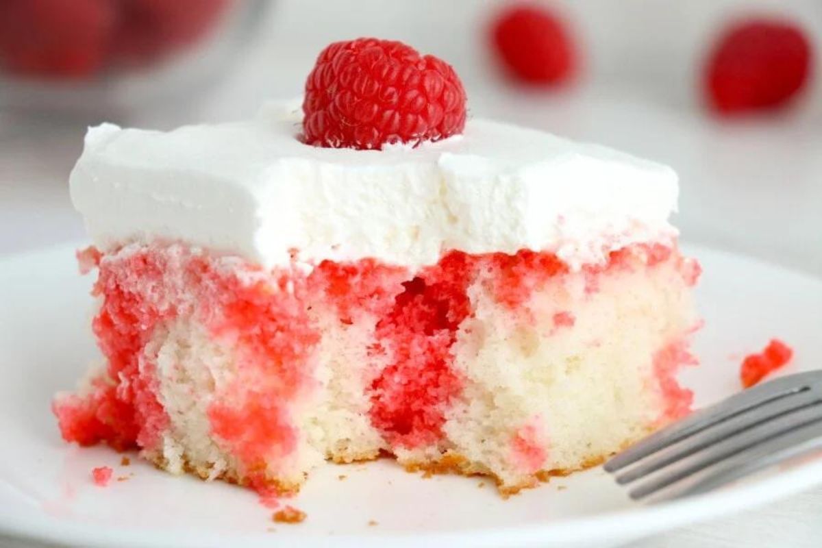 A slice of raspberry swirl cake topped with whipped cream and a fresh raspberry on a white plate, inspired by Jello recipes.