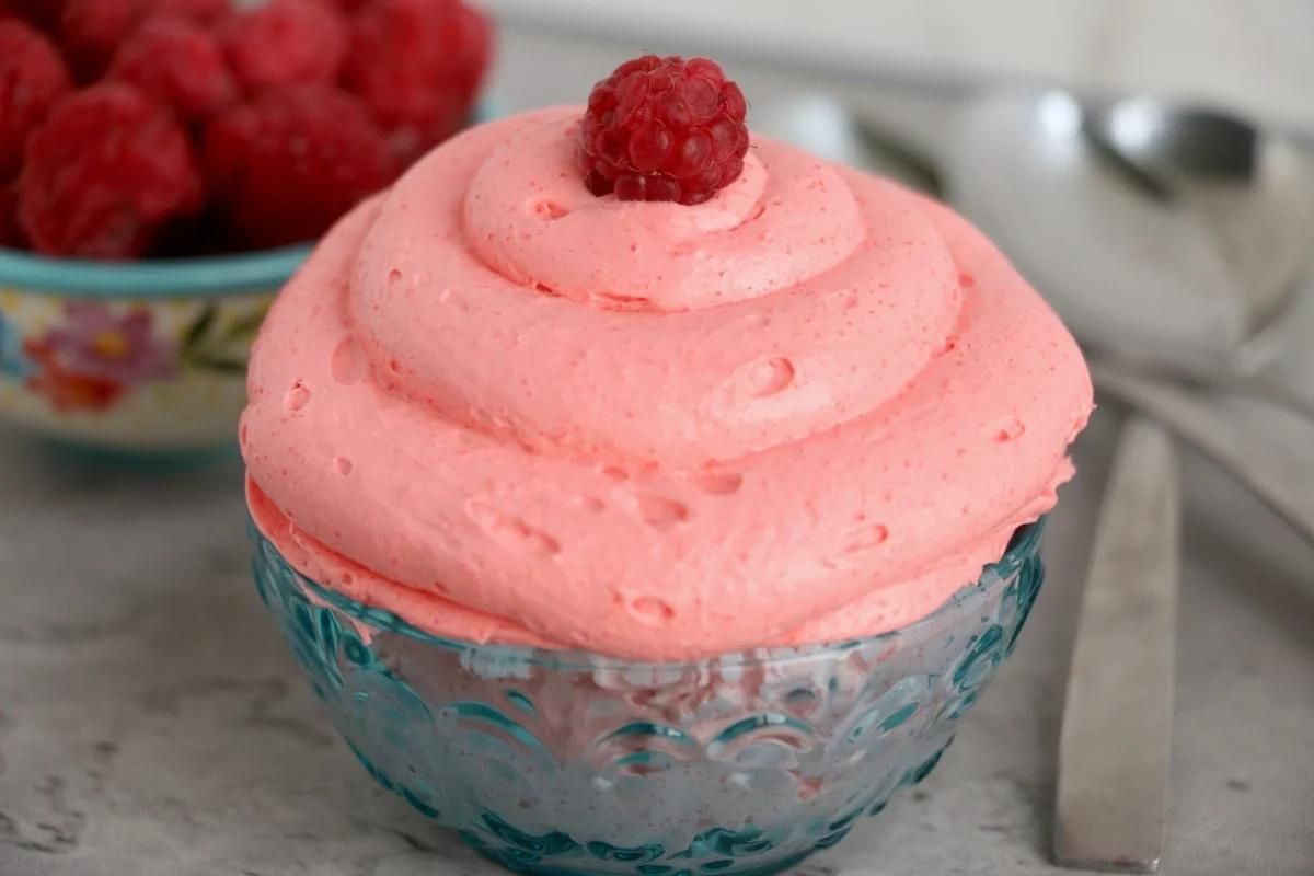 A serving of pink raspberry Jello mousse topped with a single raspberry in a decorative glass bowl.