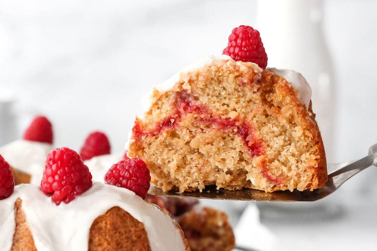 A slice of Easter raspberry cake with white frosting, served on a cake server with raspberries on top.