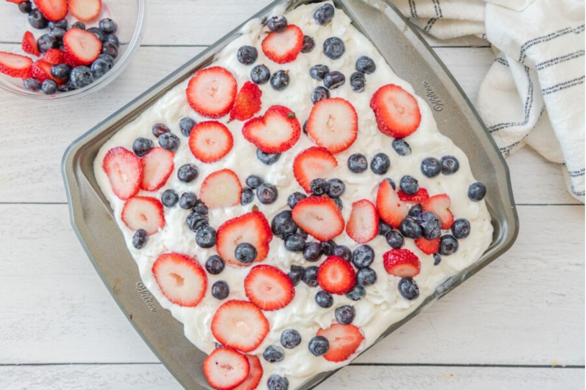 A dessert tray with Poke Cake and cupcakes topped with whipped cream, sliced strawberries, and blueberries.
