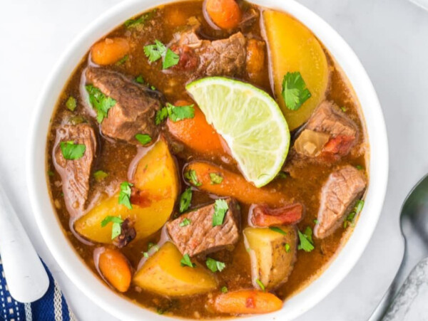 A bowl of beef stew with potatoes, carrots, herbs, and a lime wedge sits next to a spoon on a white surface—perfect inspiration for comforting stew recipes.