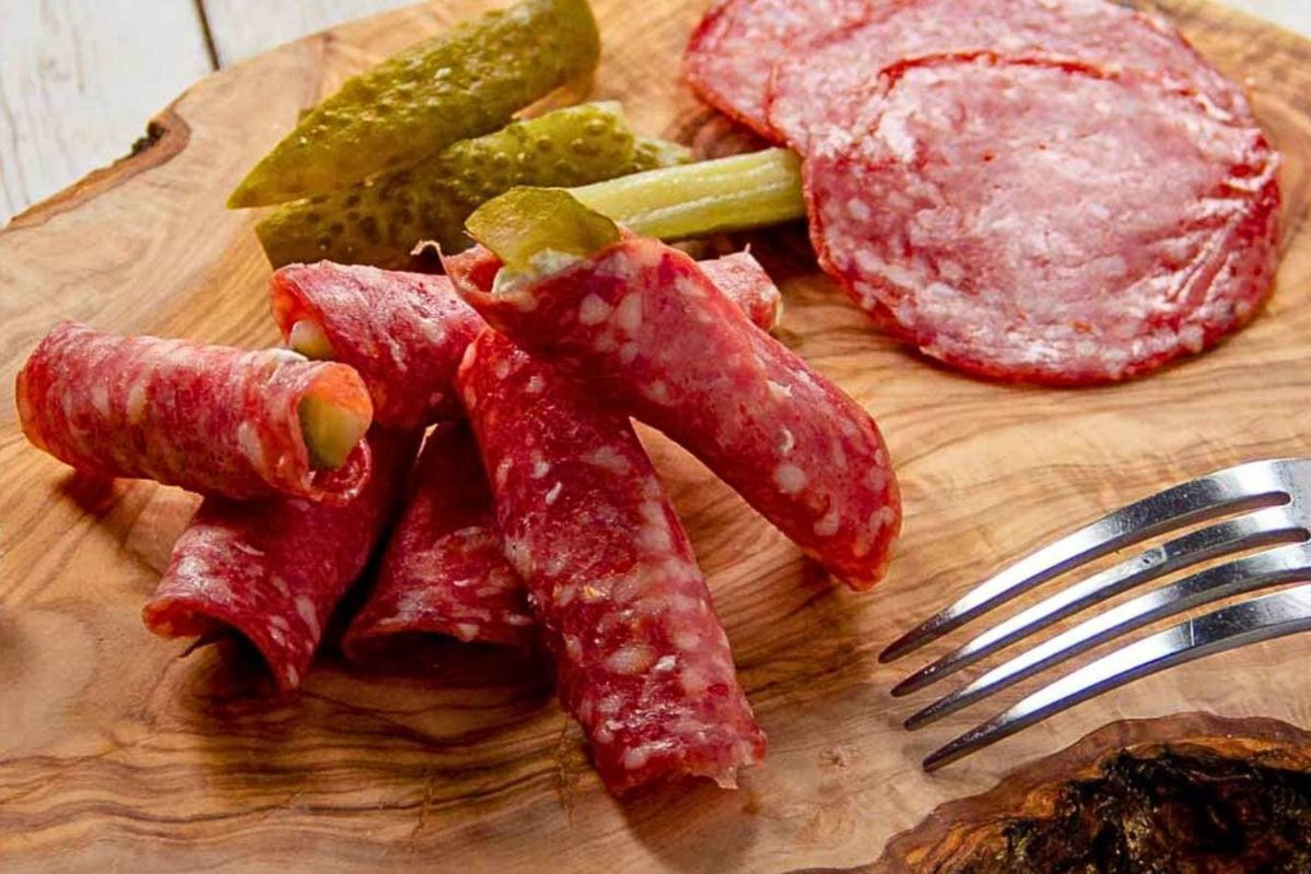 An Easter appetizer selection of sliced salami and cheese rolls with pickles on a wooden cutting board, accompanied by a fork.