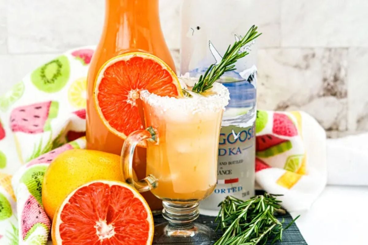 An easter citrus cocktail garnished with a grapefruit slice and rosemary, beside a bottle of vodka and fruit slices.