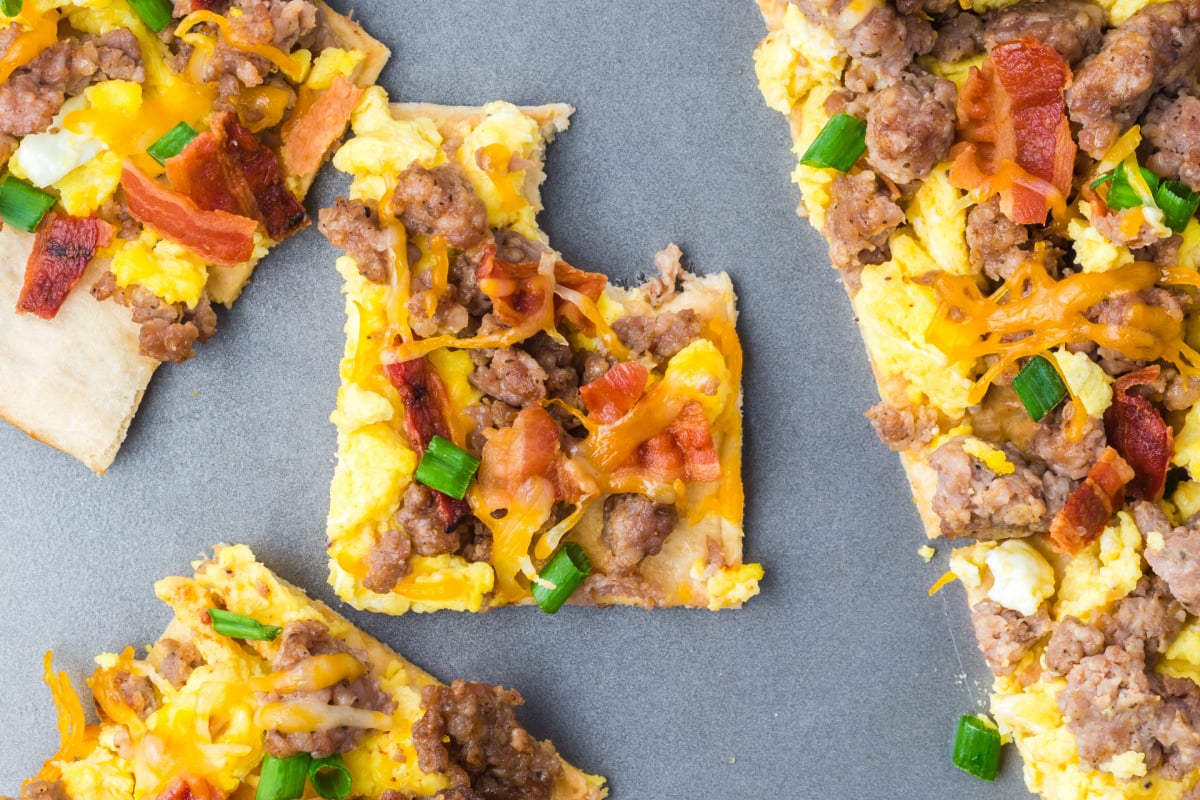 Pieces of an old school recipes breakfast pizza with scrambled eggs, sausage, cheese, and vegetables on a gray surface.