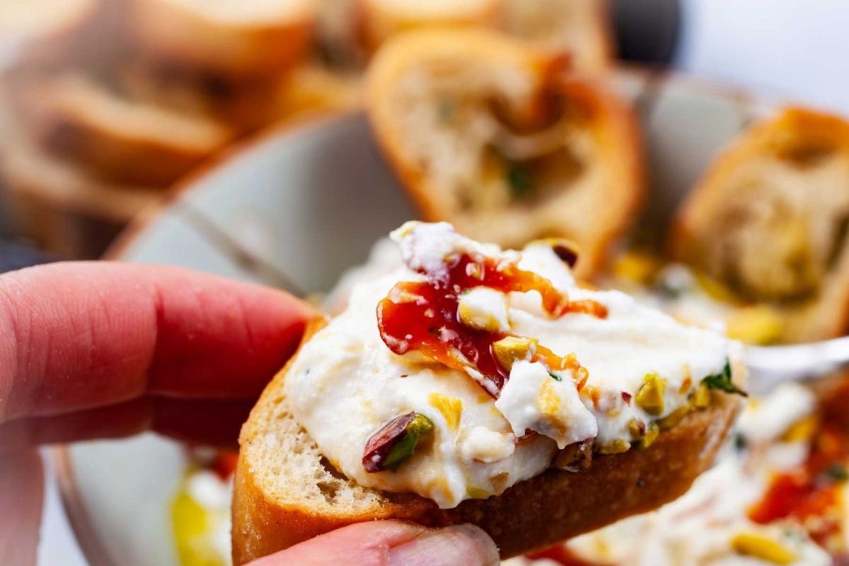 A close-up of a hand holding a piece of baguette topped with cream cheese, herbs, and a drizzle of sauce from our cheesy recipes collection.