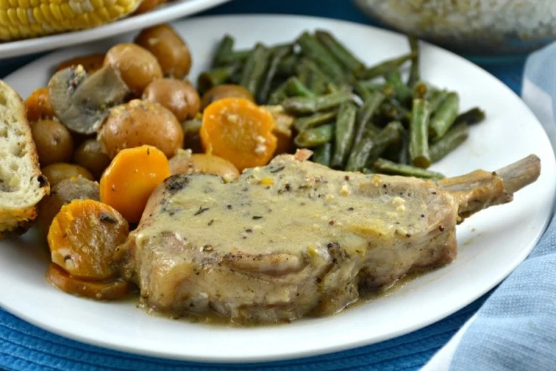 A plate of dishes with potatoes, consisting of a pork chop with gravy, green beans, baby carrots, and potatoes, with a roll on the side.