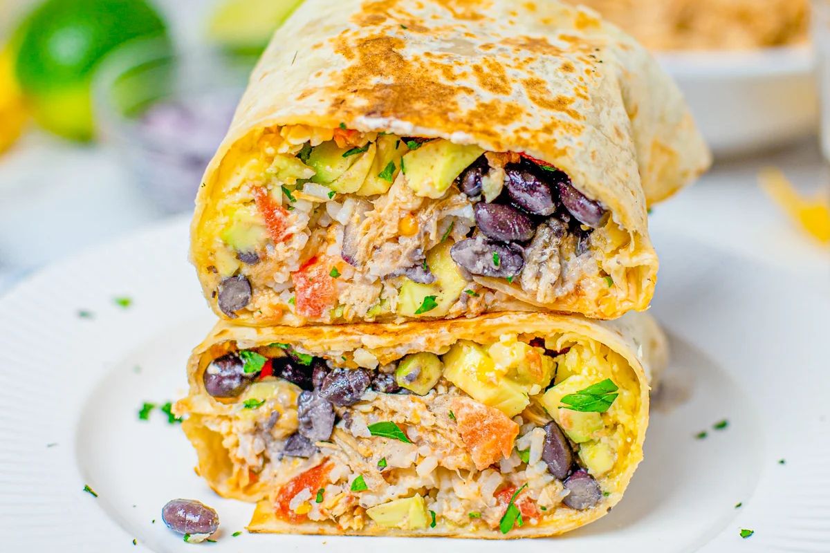 A close-up of a burrito sliced in half, stuffed with black beans, rice, avocado, tomatoes, and cilantro, displayed on a white plate&mdash;perfect inspiration for new burrito recipes.