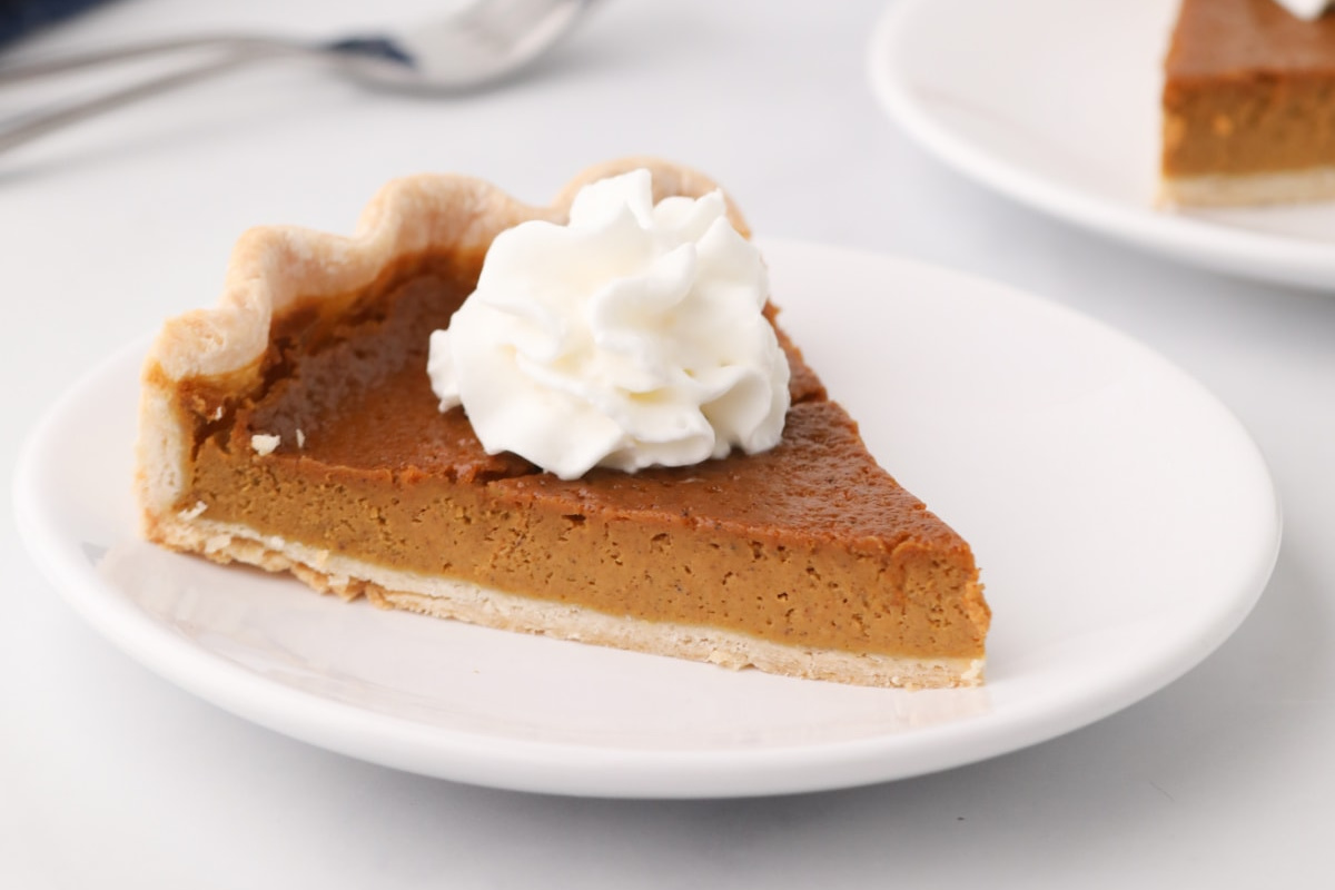A slice of pumpkin pie topped with whipped cream on a white plate, featuring in our pie recipes.