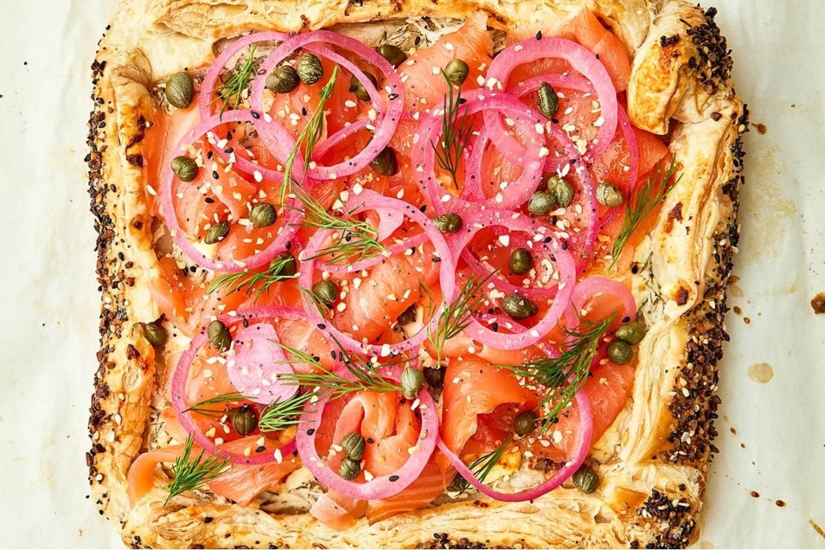 A cheesy puff pastry topped with smoked salmon, capers, pickled red onions, and fresh dill, sprinkled with sesame seeds.