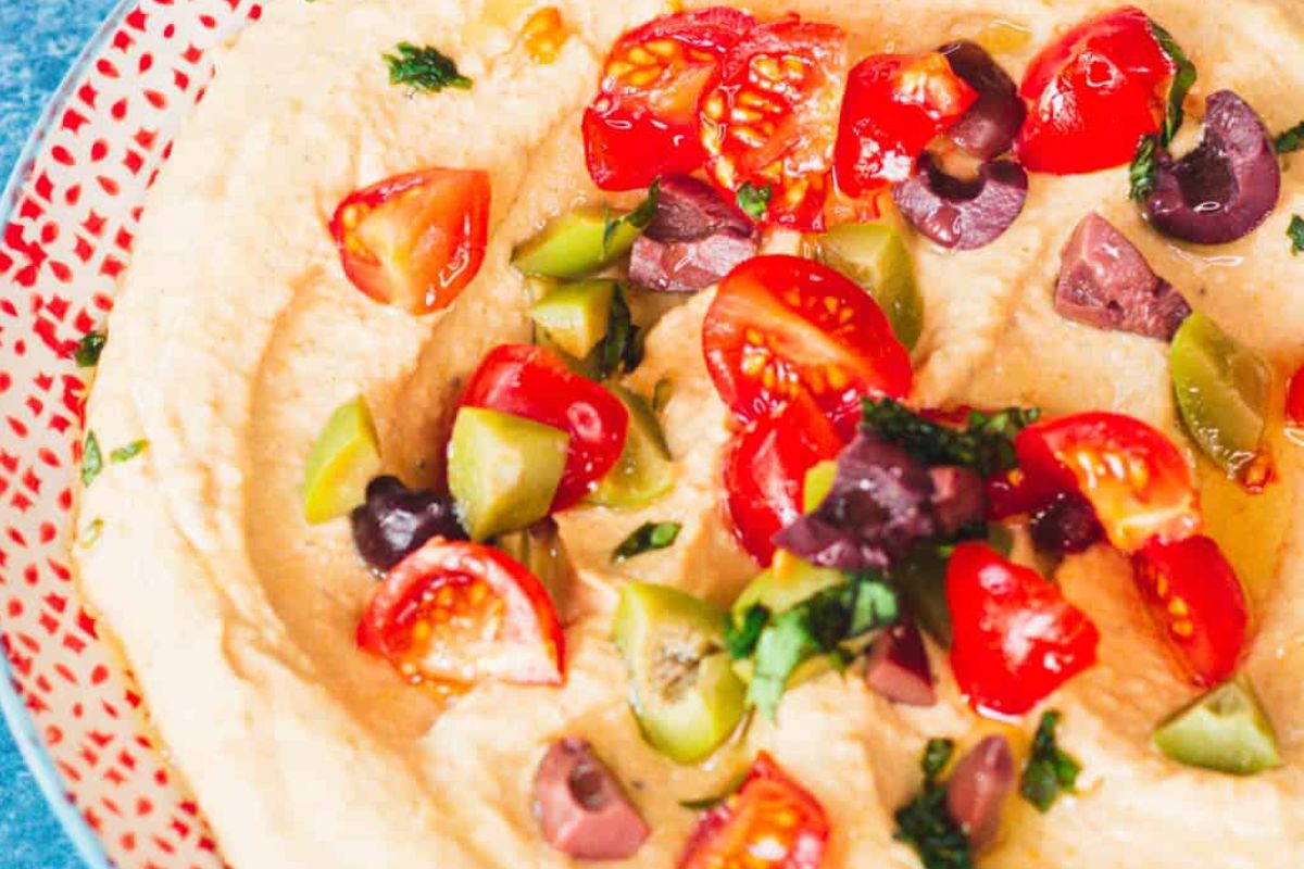 Hummus topped with tomatoes, olives, and herbs served on a patterned plate as an Easter appetizer.