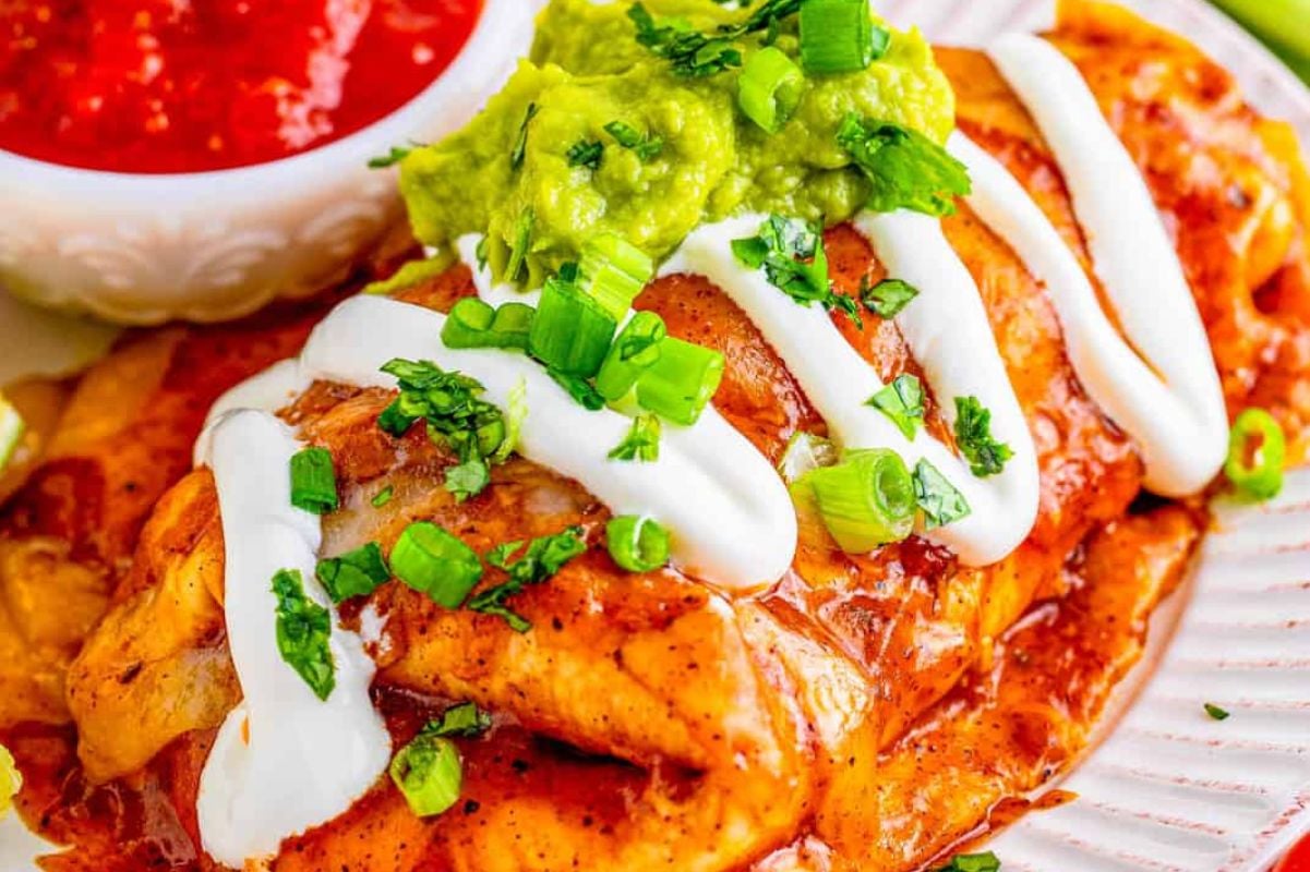 A plate of enchiladas topped with sour cream, guacamole, and fresh green onions, served with a side of salsa and burrito recipes.