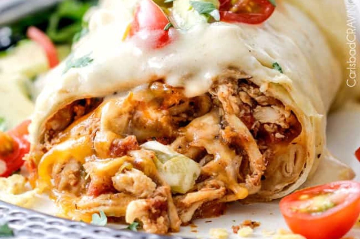 A close-up of a sliced chicken burrito recipe topped with melted cheese and garnished with diced tomatoes and herbs, served on a white plate.