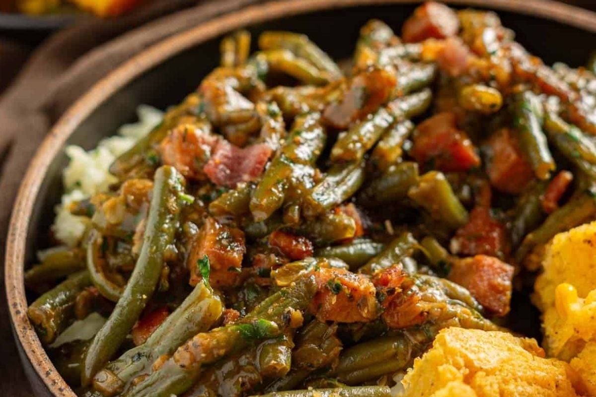 A bowl of cooked green beans mixed with diced bacon and seasonings, accompanied by cornbread on the side.