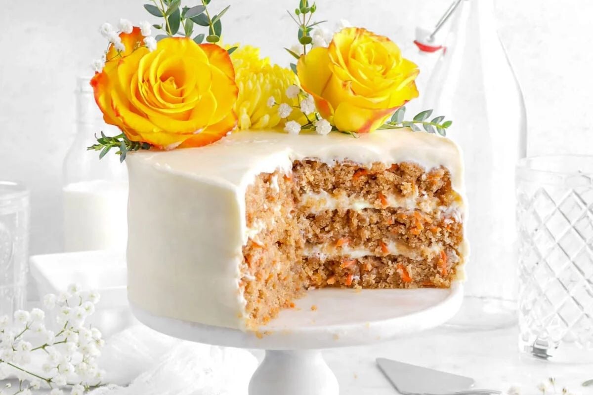 Three-layer carrot cake with white frosting, topped with yellow roses and greenery, missing one slice—a perfect choice for Easter cakes.