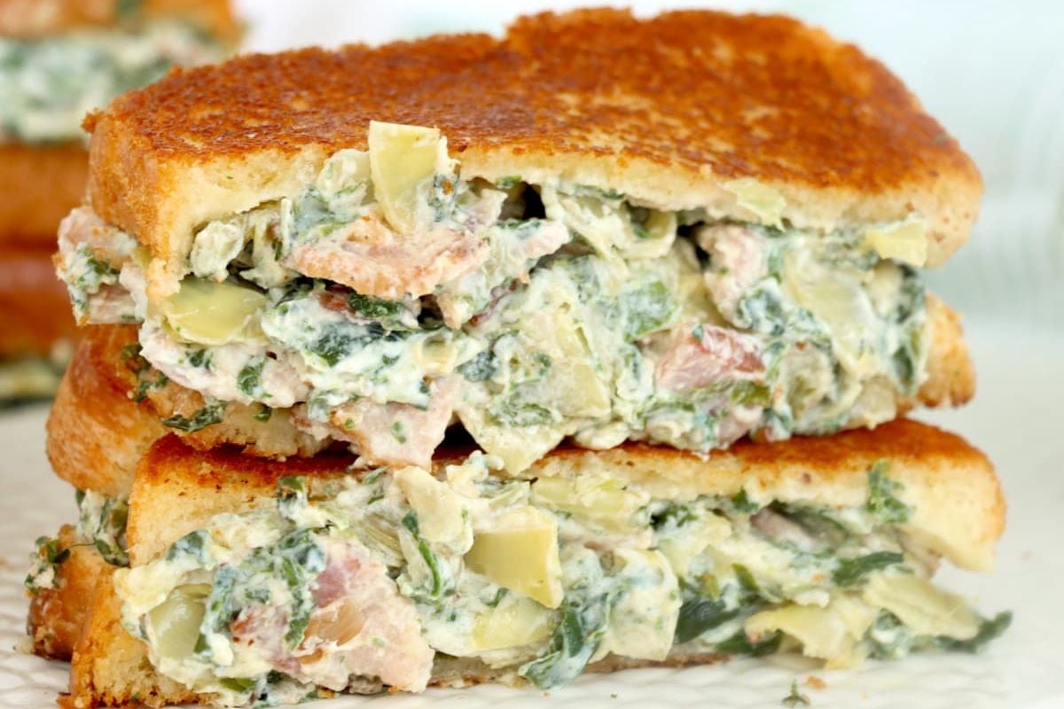 A close-up of a toasted sandwich filled with a creamy, cheesy spinach and artichoke dip, with visible chunks of artichoke.