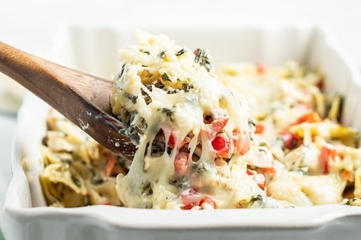 A wooden spoon is lifting a serving of cheesy baked pasta from a white casserole dish, featuring cheesy recipes with spinach and tomatoes.