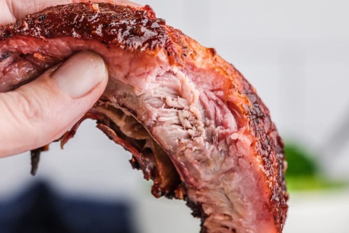 A close-up of a hand holding a piece of juicy, cooked pork rib from an easy rib recipe.